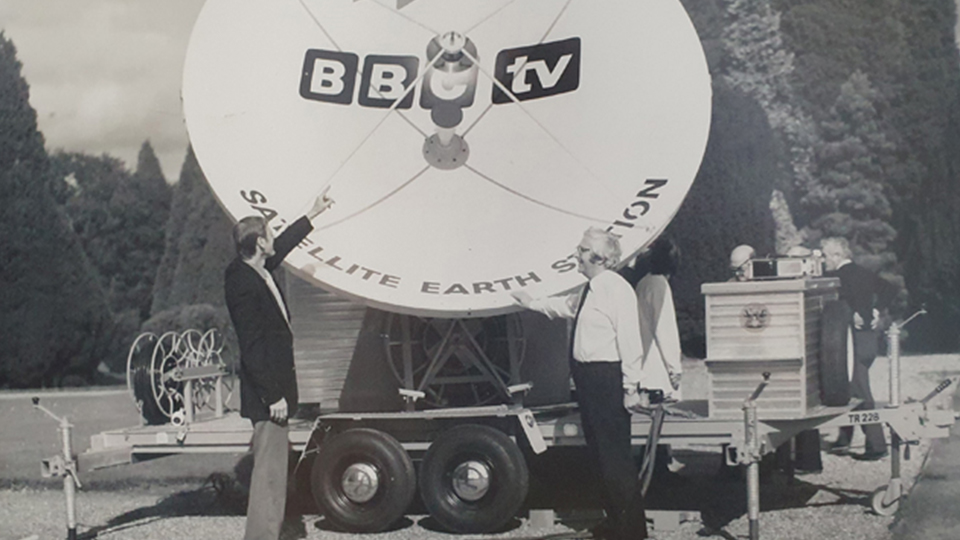 Two people stand in front of a large satellite dish which is on a wheeled truck. The BBC logo in on the satellite dish. 